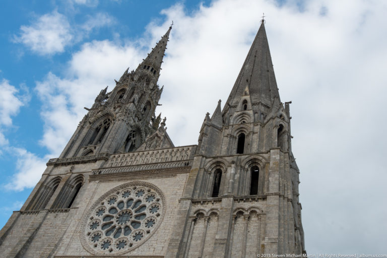 Low-angle view of Chartres Cathedral’s ornate north spire, simpler south spire, and rose window against a cloudy sky.