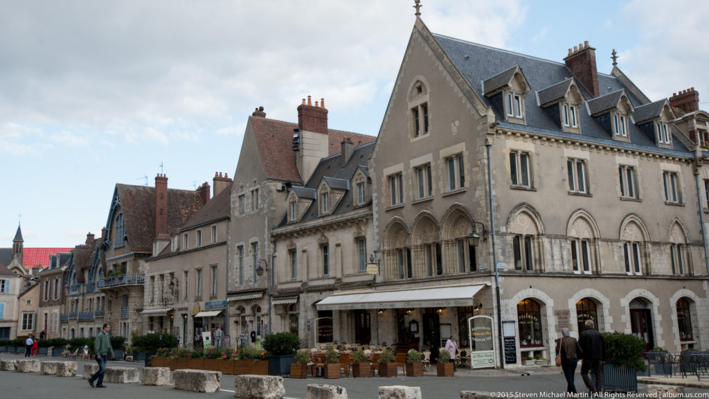 Historic stone townhouses with steep roofs, dormers, shops, café seating, and pedestrians in central Chartres.