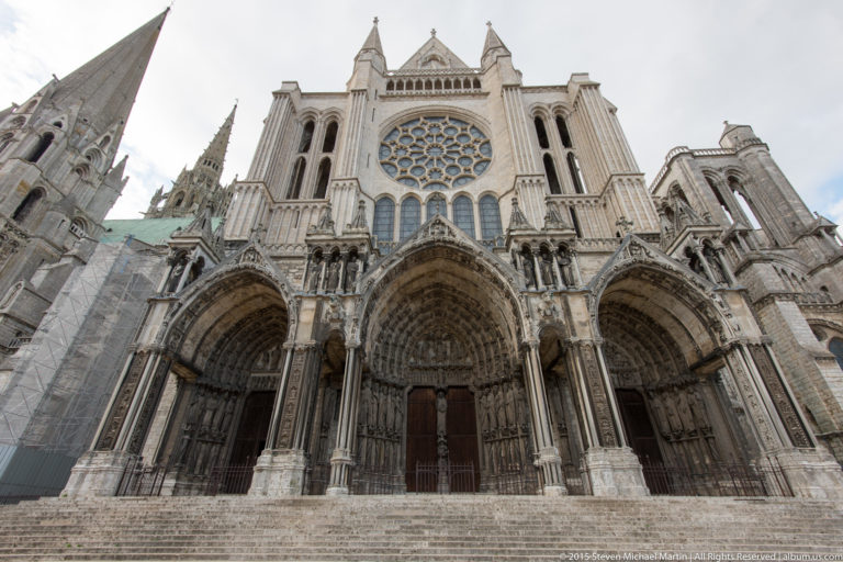 Low-angle view of Chartres Cathedral’s north transept façade with three sculpted portals, broad steps, lancets, and a rose window.