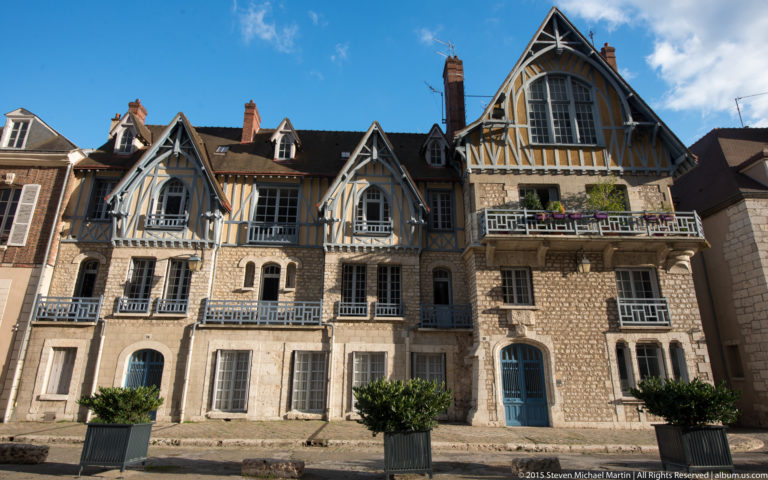 Sunlit historic building with blue-and-ochre timber-framed gables, stone lower stories, balconies, and clipped street trees in Chartres.