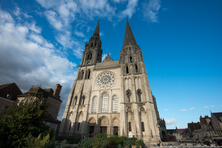 Sunlit west façade of Chartres Cathedral with contrasting twin spires and central rose window beneath a vivid blue sky.