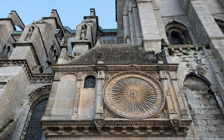 Large circular historic clock with radiating metal spokes and numbered ring mounted on the stone exterior of Chartres Cathedral.