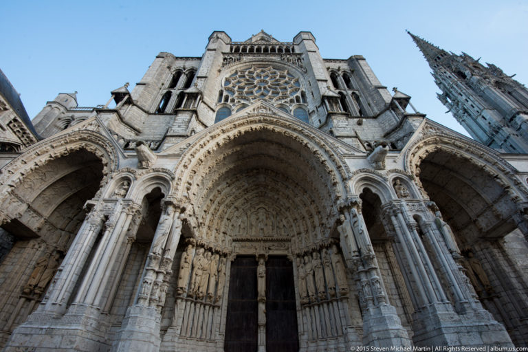 Steep low-angle view of Chartres Cathedral’s north portal with sculpted archivolts, jamb figures, rose window, and spire.