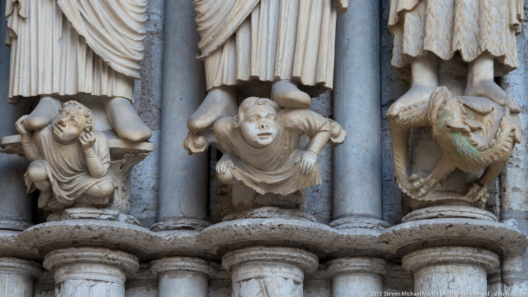Close view of small carved human figures and a fantastical creature beneath larger stone portal statues at Chartres Cathedral.