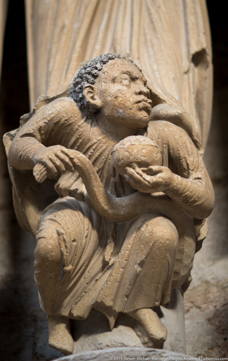 Close-up of a crouching carved stone figure beneath the drapery of a larger portal statue at Chartres Cathedral.