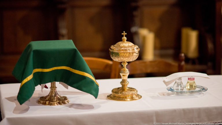 Ornate gold-colored liturgical vessel with a cross beside a green-covered object and smaller vessels on a white altar cloth.