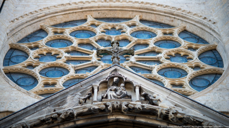 Stone rose-window tracery above a steep sculpted gable with a small crucifix on the exterior of Chartres Cathedral.