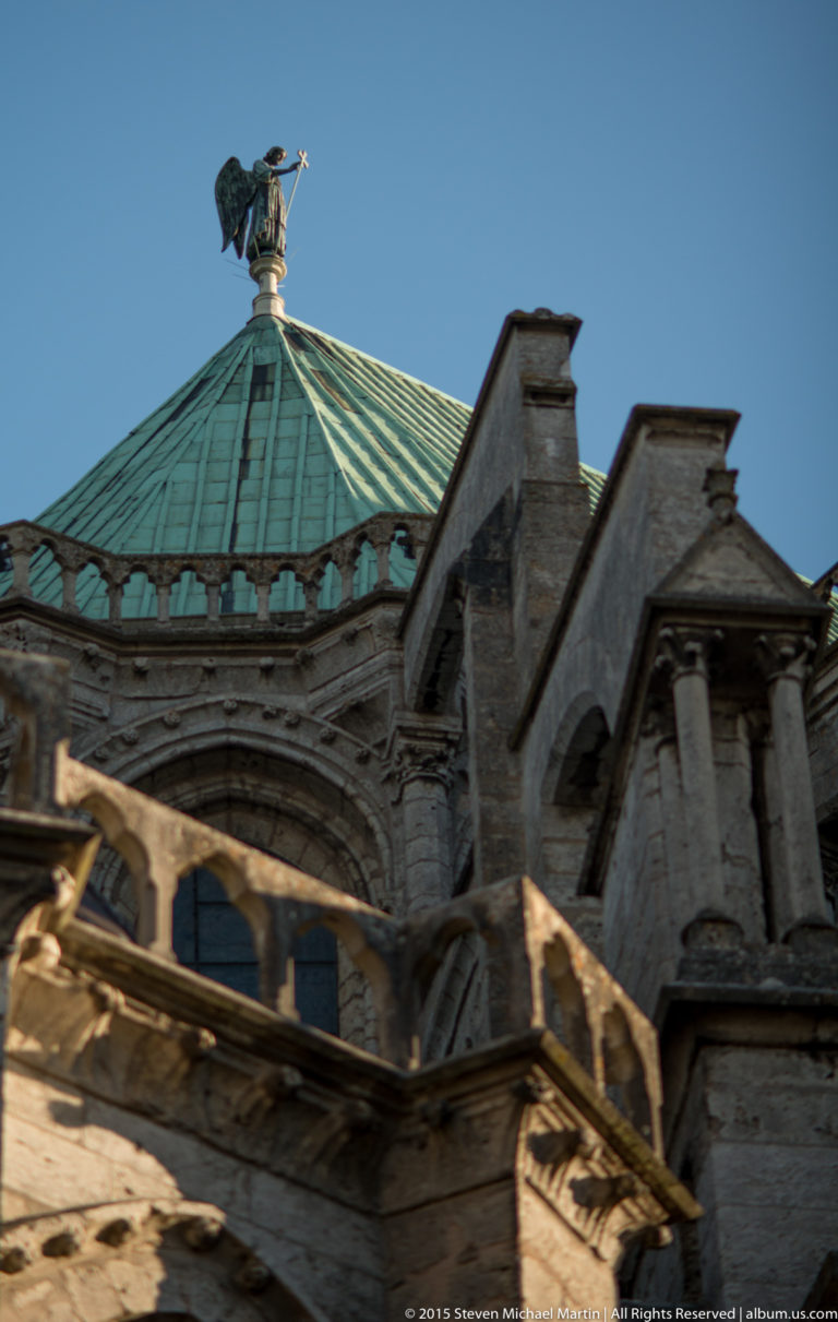 Green-patinated polygonal roof rising above layered stone buttresses and masonry on Chartres Cathedral under a blue sky.