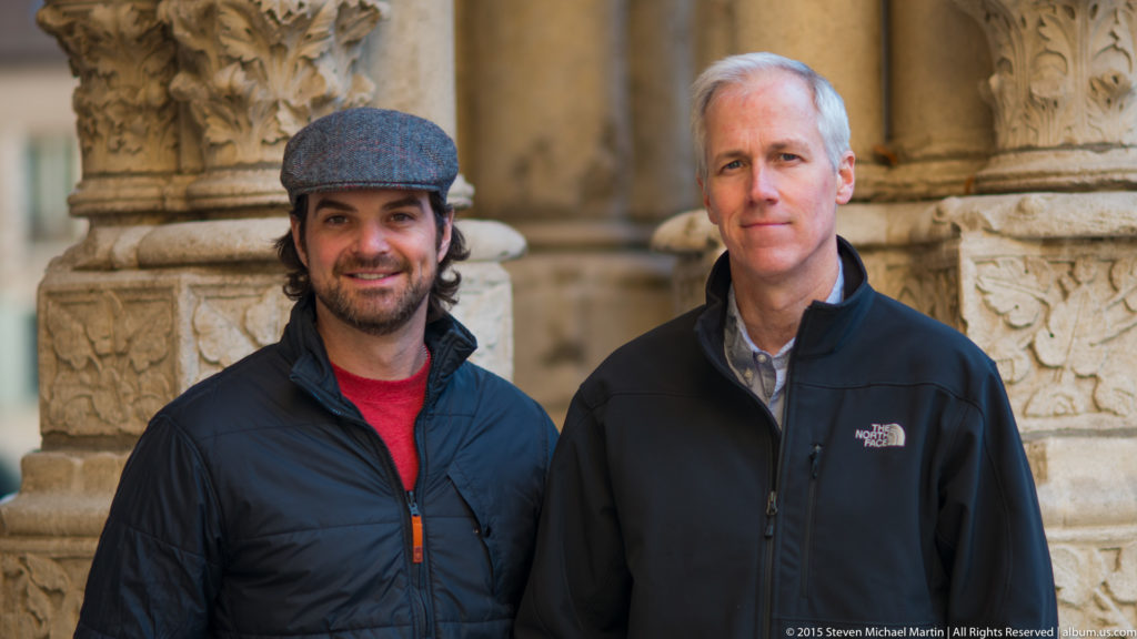 Two men, one wearing a flat cap, standing together before carved stone columns at Chartres Cathedral.