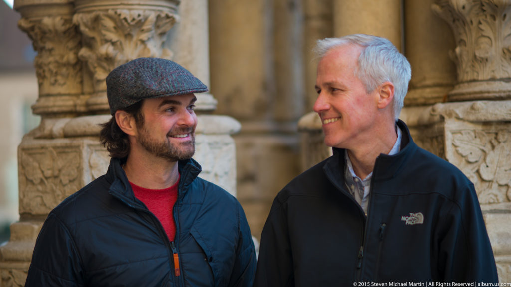 Two men smiling toward each other in front of carved stone columns at Chartres Cathedral, one wearing a flat cap.