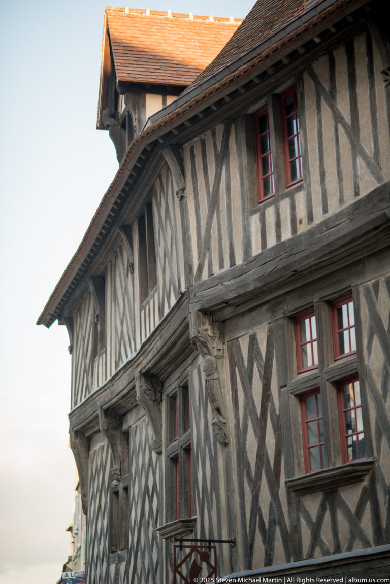 Close view of a half-timbered house with dark diagonal beams, pale infill, red-trimmed windows, and projecting tiled roof in Chartres.