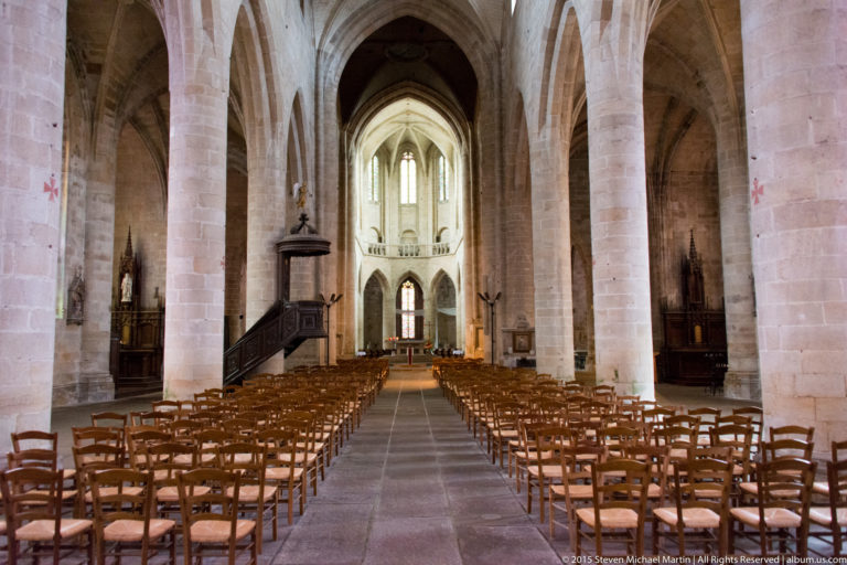 Central aisle of Église Saint-Malo with wooden chairs, pale stone piers, pointed arches, ribbed vaults, pulpit, and distant choir.