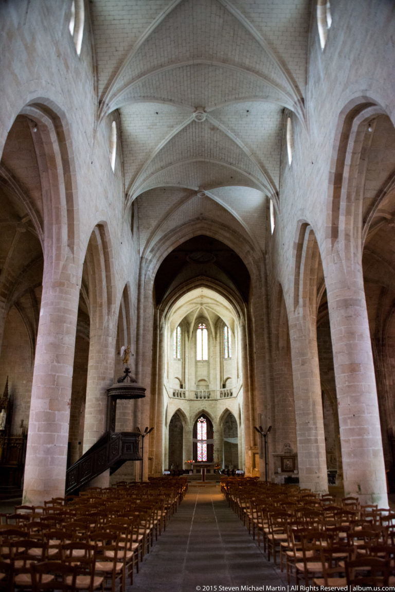 Wide interior view of Église Saint-Malo showing ribbed vaults, pointed arcades, stone piers, rows of chairs, and a carved pulpit.