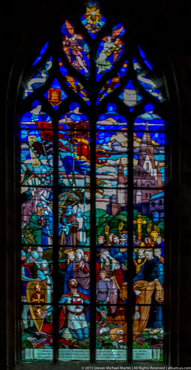 Tall multicolored stained-glass window with figurative scenes, heraldic motifs, and Gothic tracery inside Église Saint-Malo.