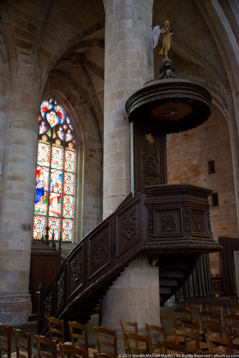Dark carved wooden pulpit with enclosed stair attached to a granite pier beside a stained-glass window in Église Saint-Malo.