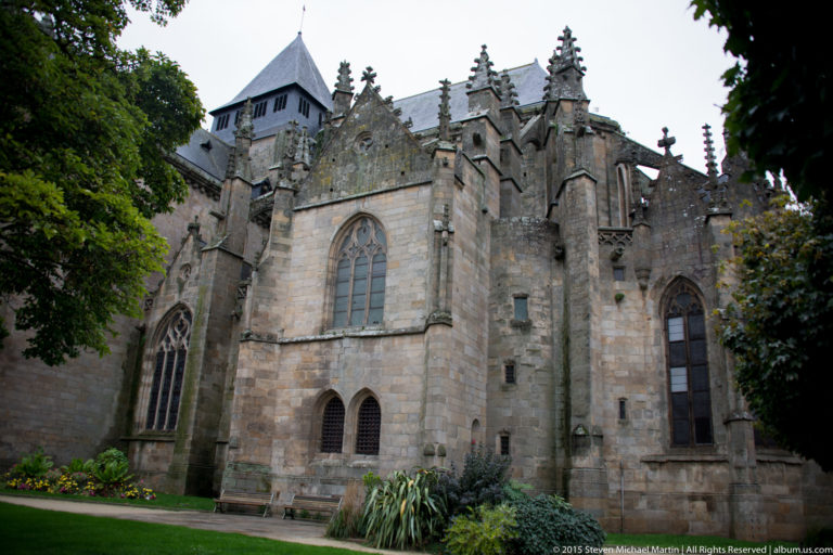 Oblique exterior view of Église Saint-Malo with tall Gothic windows, buttresses, pinnacles, slate roofs, and a small lawn.