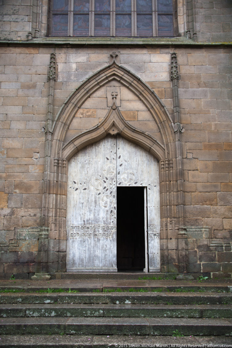Large pointed stone portal with weathered doors, one leaf partly open, on the exterior of Église Saint-Malo in Dinan.