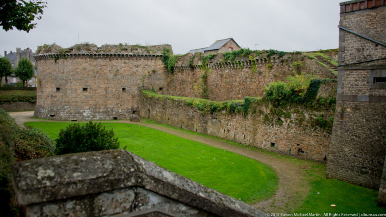 Round stone defensive tower joined to a long curtain wall above a grassy ditch and path in Dinan.