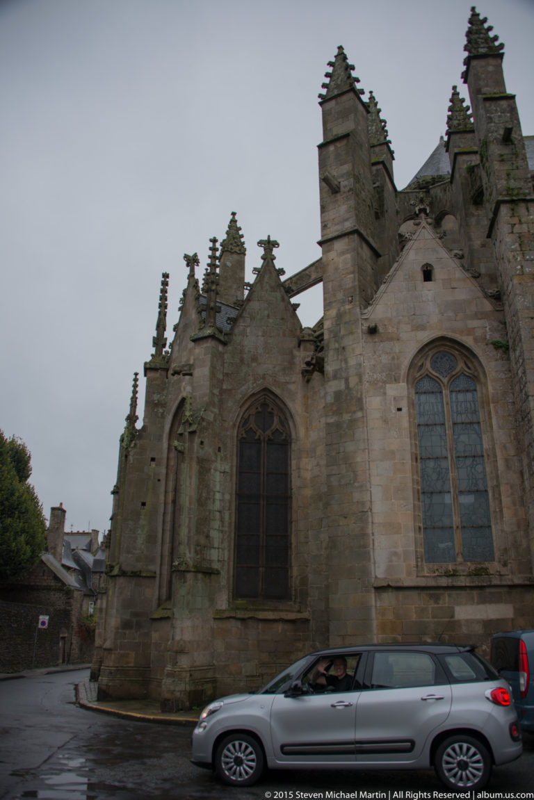 Street-level side view of Église Saint-Malo with Gothic windows, buttresses, steep roof, and cars passing in front.