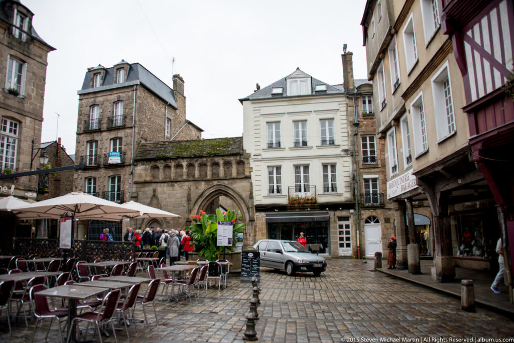 Cobbled square with café seating, historic façades, and a pointed stone passage in the center of Dinan.