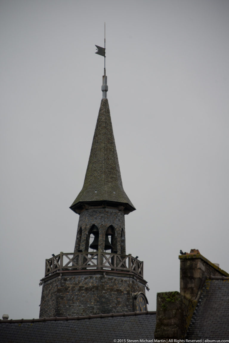Upper section of Dinan’s Tour de l’Horloge with stone belfry, open balustrade, steep slate roof, and weather vane.