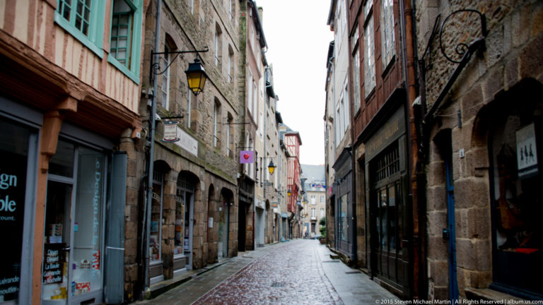 Narrow wet cobblestone street lined by old stone and timber-fronted shops in the historic center of Dinan.