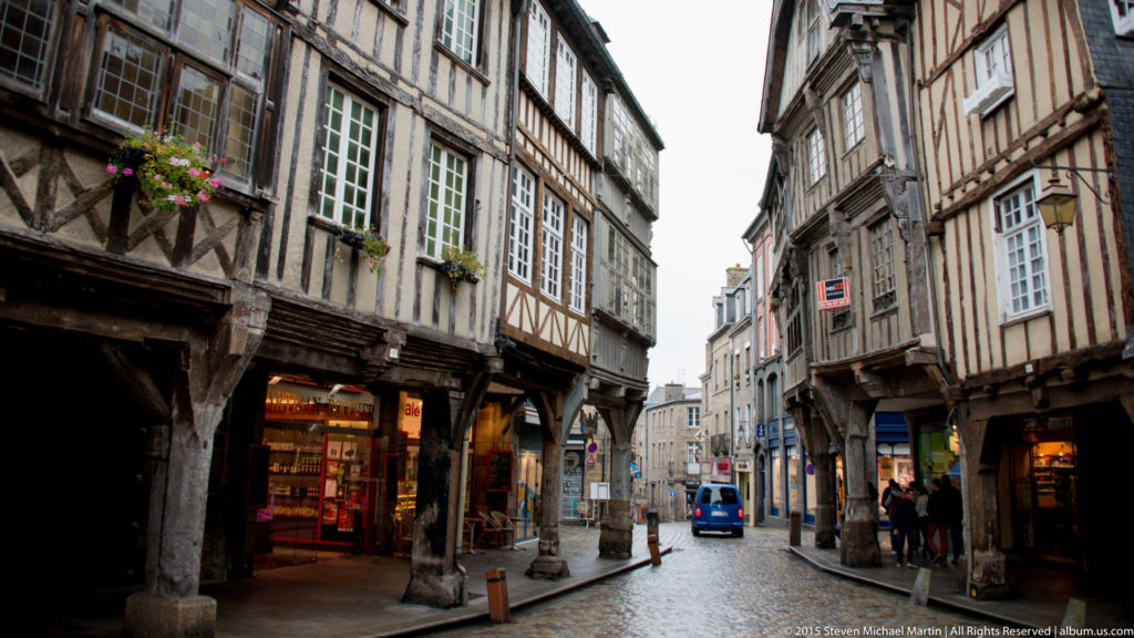 Historic Dinan street with projecting half-timbered houses, exposed posts and braces, shopfronts, and wet cobbles.