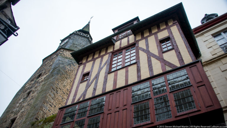 Upward view of a red-brown half-timbered house with the Tour de l’Horloge rising behind it in Dinan.
