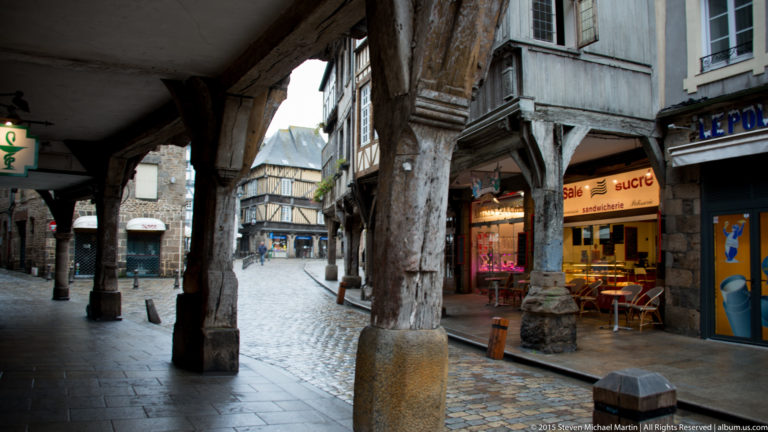 View beneath heavy timber posts and overhanging upper floors along a cobbled commercial street in historic Dinan.