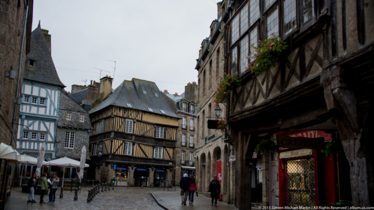 Cobbled Dinan street framed by dark half-timbered houses, projecting upper stories, shopfronts, and pedestrians.