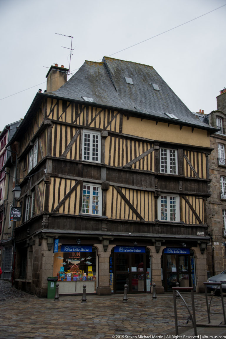 Large corner house in Dinan with dark exposed timber framing, pale infill panels, many windows, slate roof, and shops below.