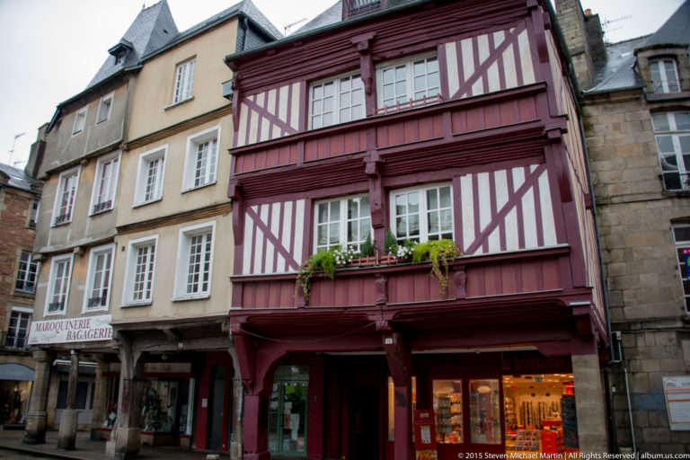 Red-and-white half-timbered house with multi-pane windows and flower boxes between stone buildings in Dinan.