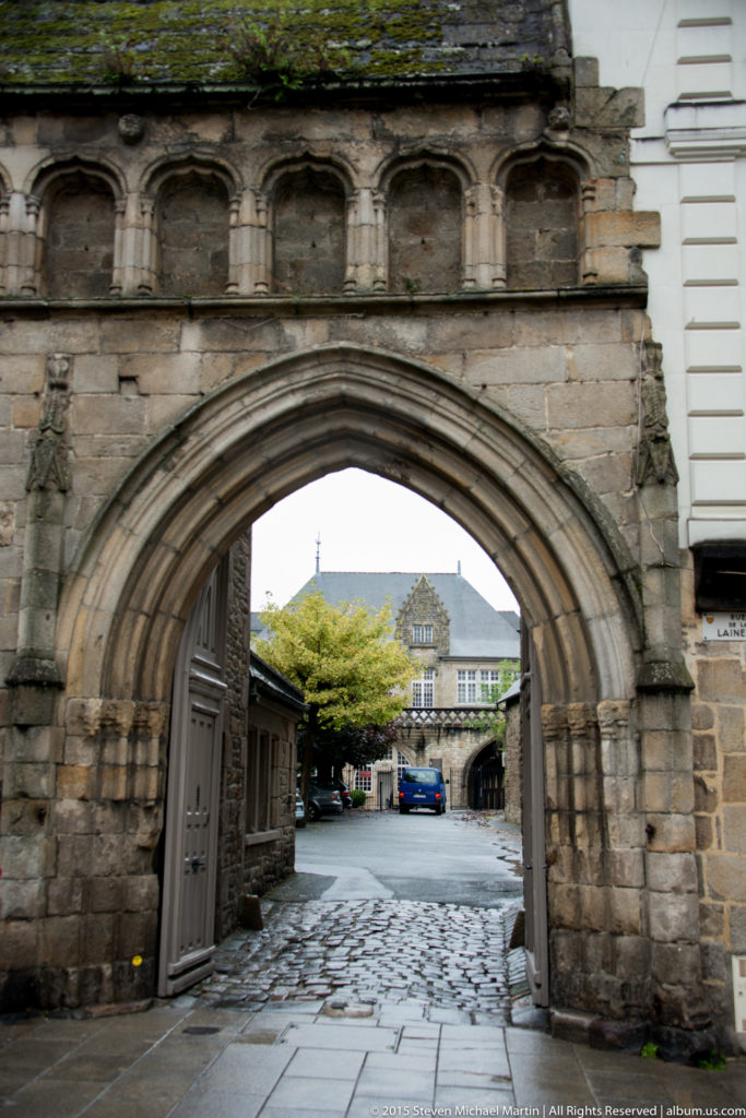 Large pointed stone archway with molded masonry and shallow niches above, opening onto a paved area in historic Dinan.
