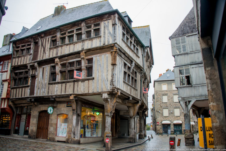 Weathered half-timbered corner house with projecting upper floors and the Tour de l’Horloge visible behind it in Dinan.