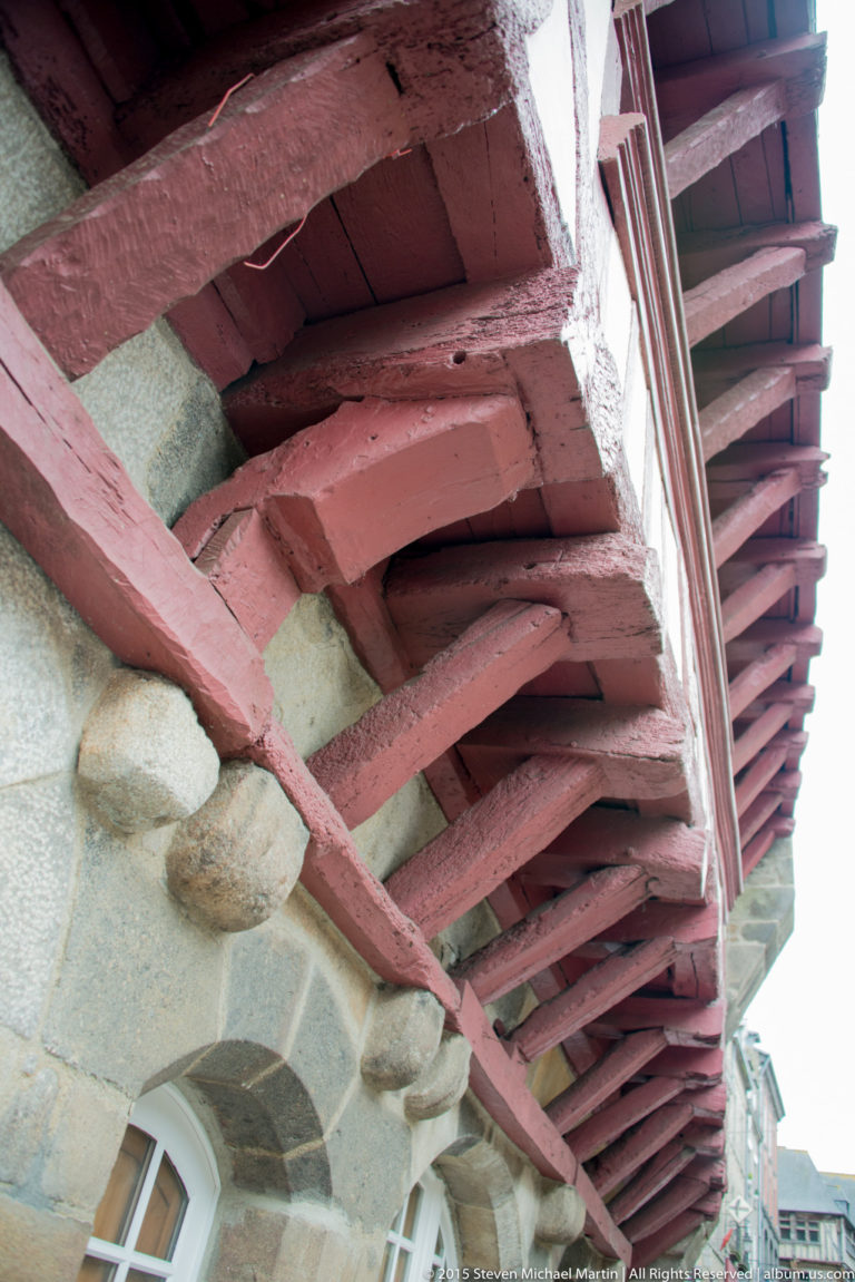 Close upward view beneath a projecting half-timbered story, showing red-painted beams, joists, braces, and stone wall below.