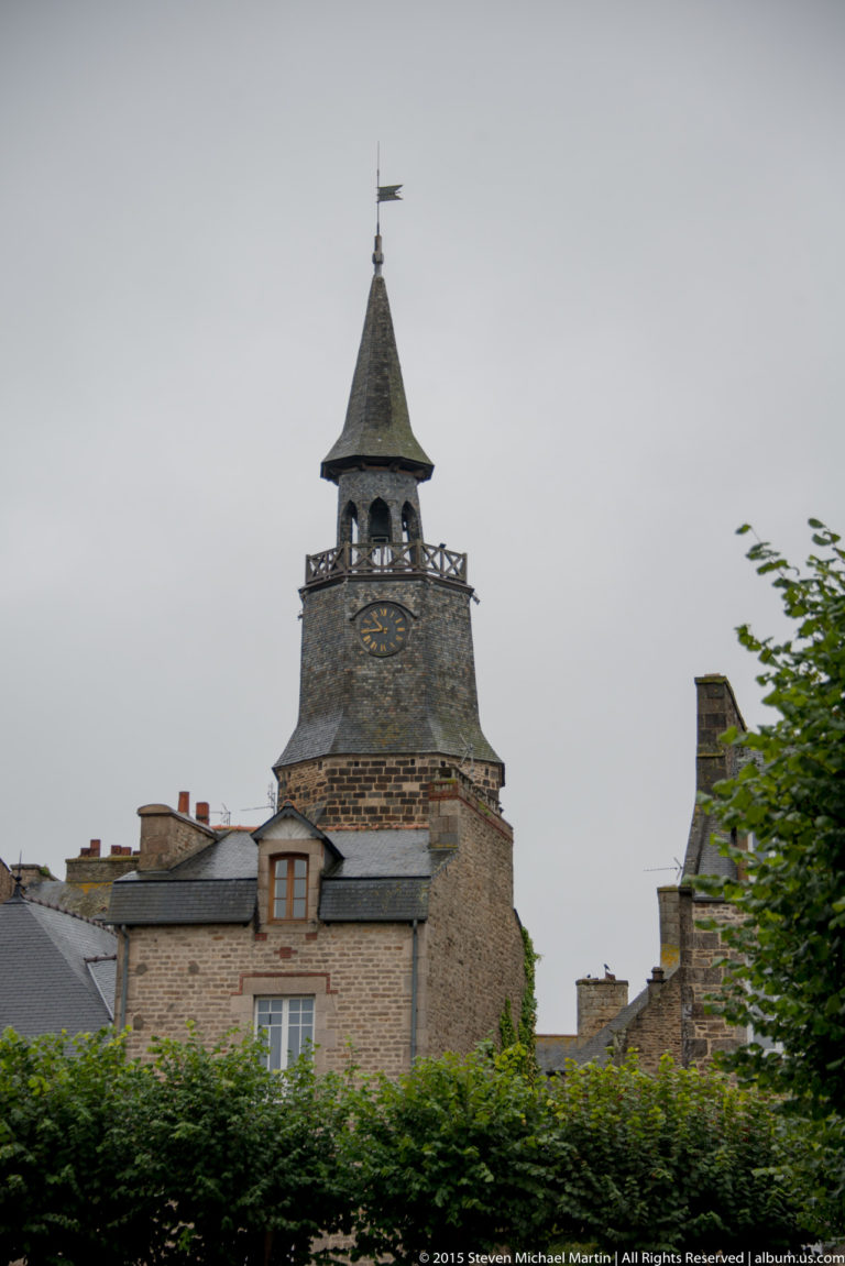 Tour de l’Horloge with visible clock face and steep slate roof rising above houses and greenery in Dinan.