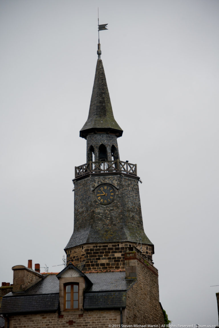Close view of Dinan’s Tour de l’Horloge showing its clock face, open belfry, balustrade, steep slate roof, and weather vane.