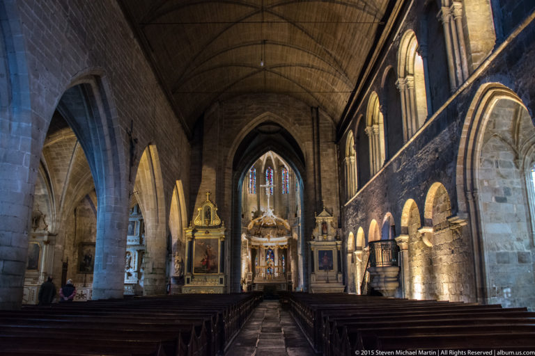 Interior of Dinan’s Basilique Saint-Sauveur with wooden pews, stone arcades, side windows, stained glass, and ornate sanctuary ahead.
