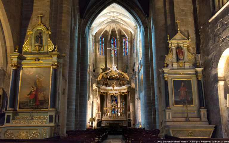 Sanctuary of the Basilique Saint-Sauveur with stained glass, gilded side altars, painted panels, stone piers, and pointed vaulting.