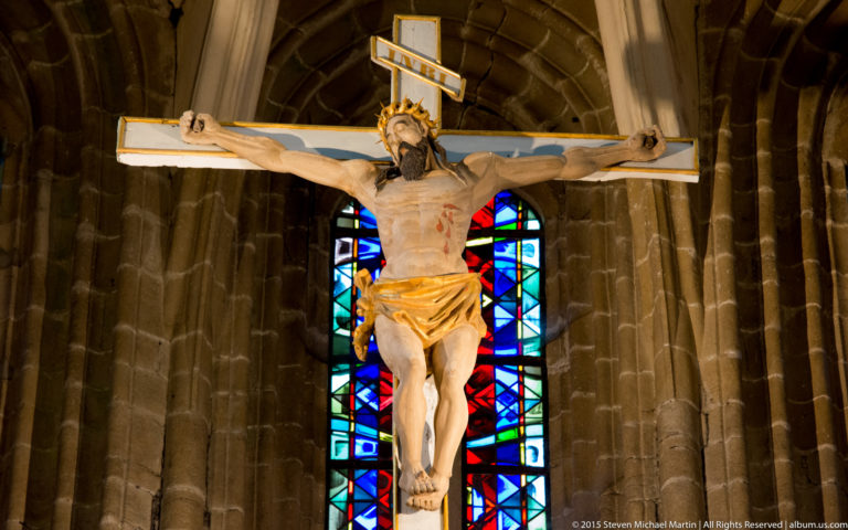 Crucifix with sculpted figure of Christ positioned before a colorful stained-glass window inside the Basilique Saint-Sauveur.