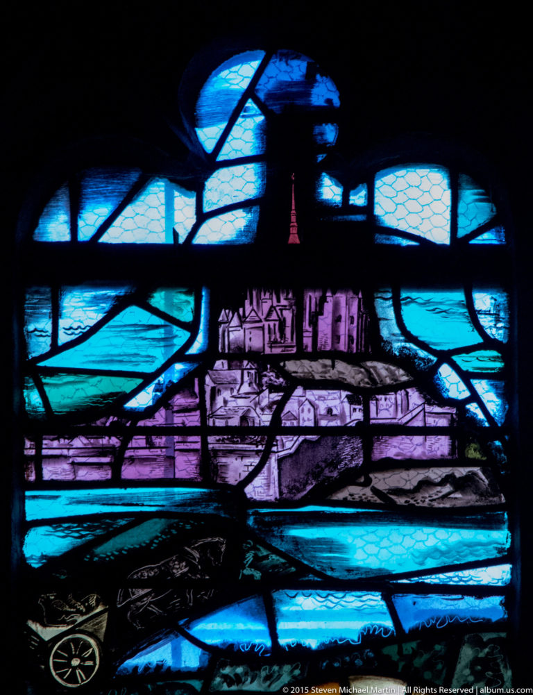 Blue and violet stained-glass panel showing a fortified settlement with towers, walls, roofs, and surrounding landscape.