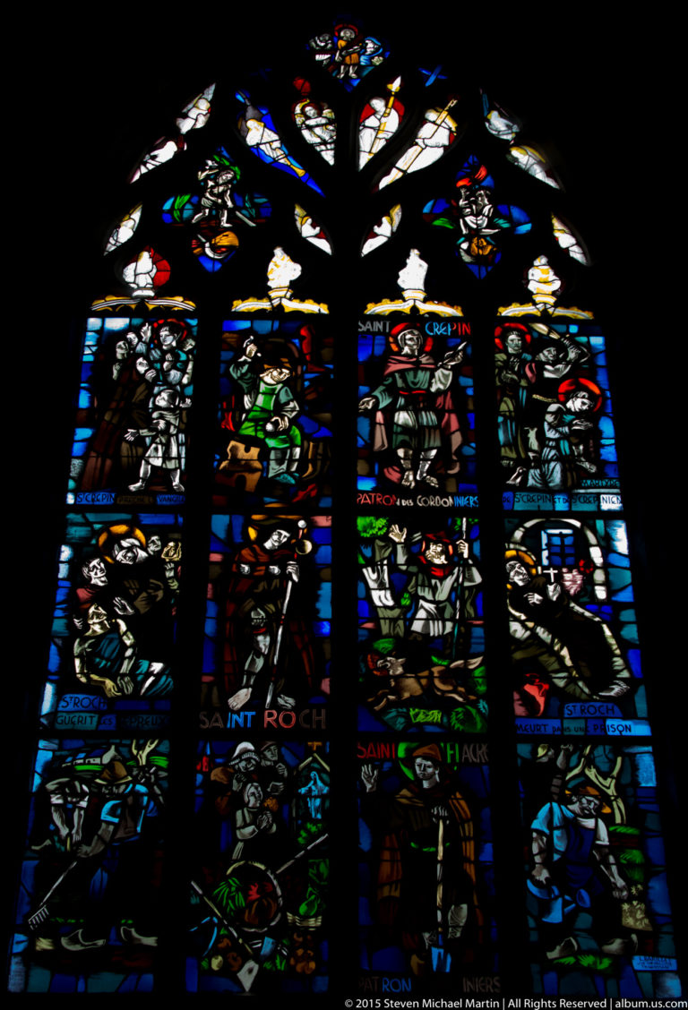 Tall stained-glass window with multiple saints, narrative panels, inscriptions, and Gothic tracery in the Basilique Saint-Sauveur.