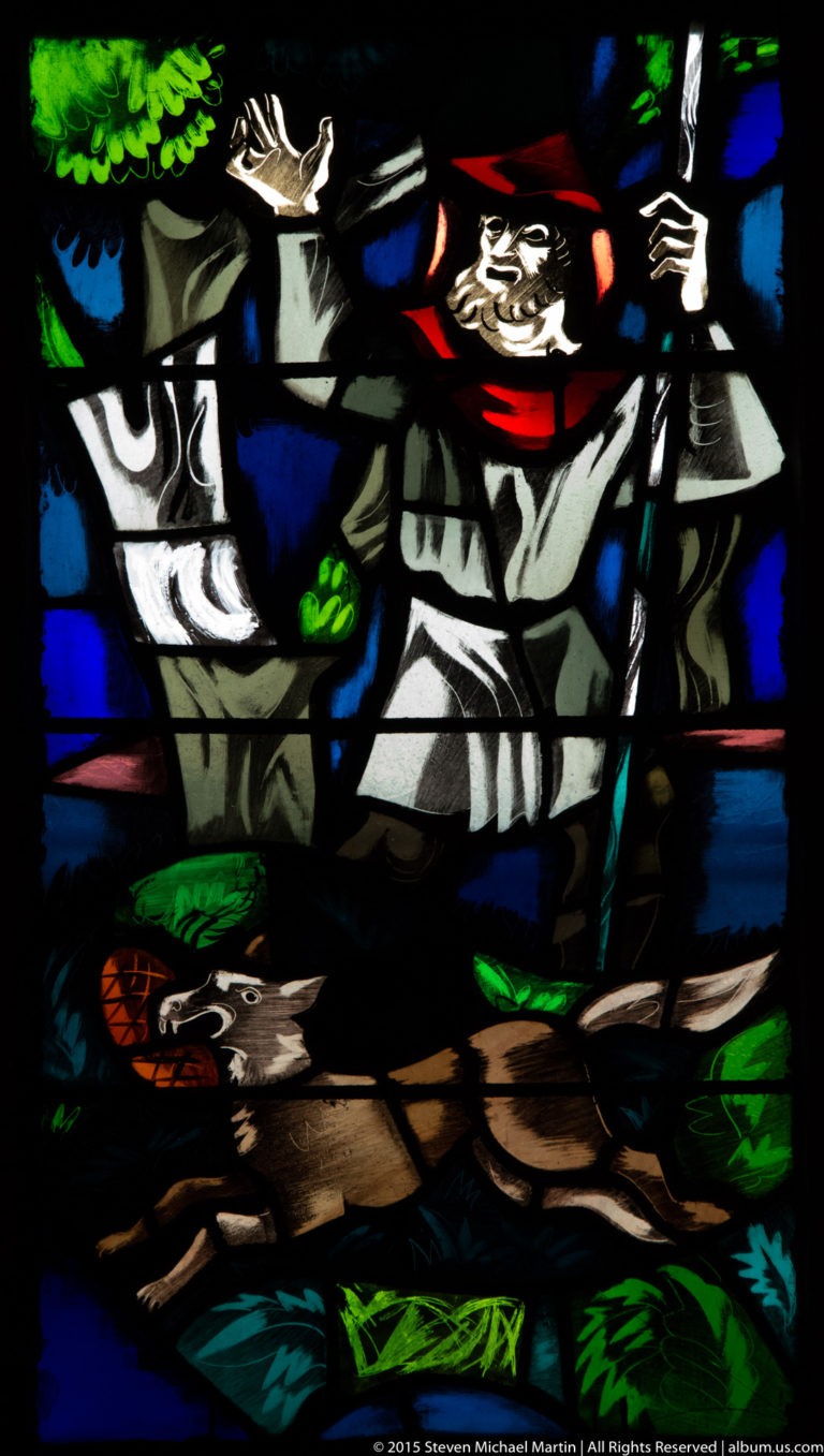 Stained-glass detail with a central robed figure raising one arm amid other figures and colored landscape elements.
