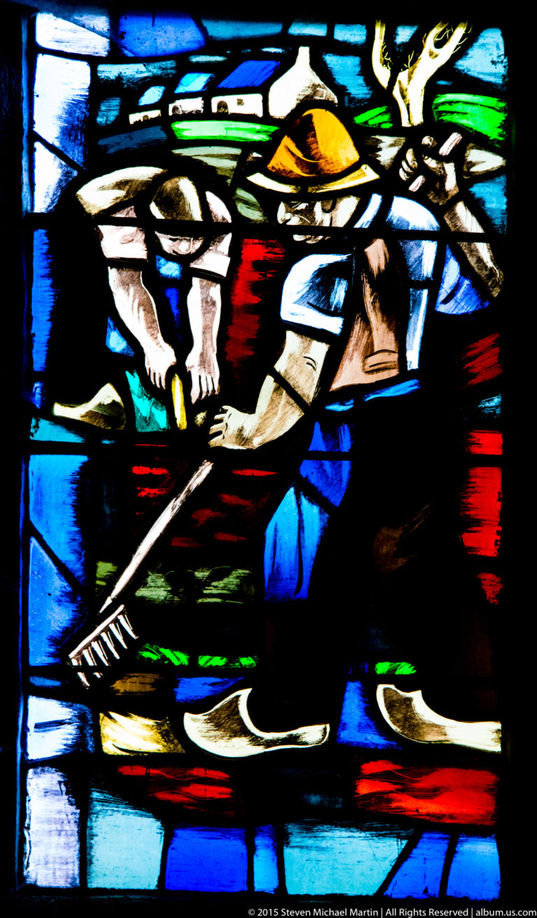 Stained-glass scene with one kneeling figure working near the ground and another standing behind in a bright landscape.