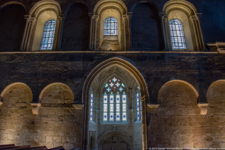 Stone interior elevation with heavy round arches, upper windows, a pointed central opening, and stained glass in the Basilique Saint-Sauveur.