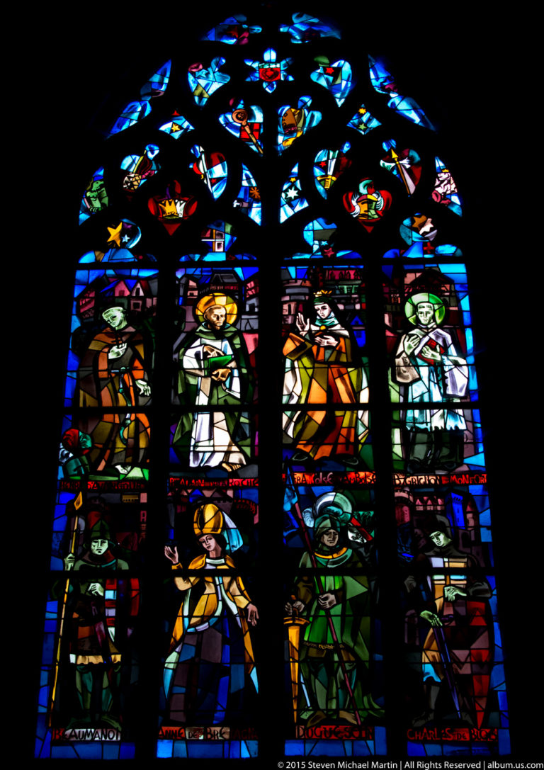 Tall stained-glass window with standing figures and narrative scenes beneath elaborate tracery in the Basilique Saint-Sauveur.
