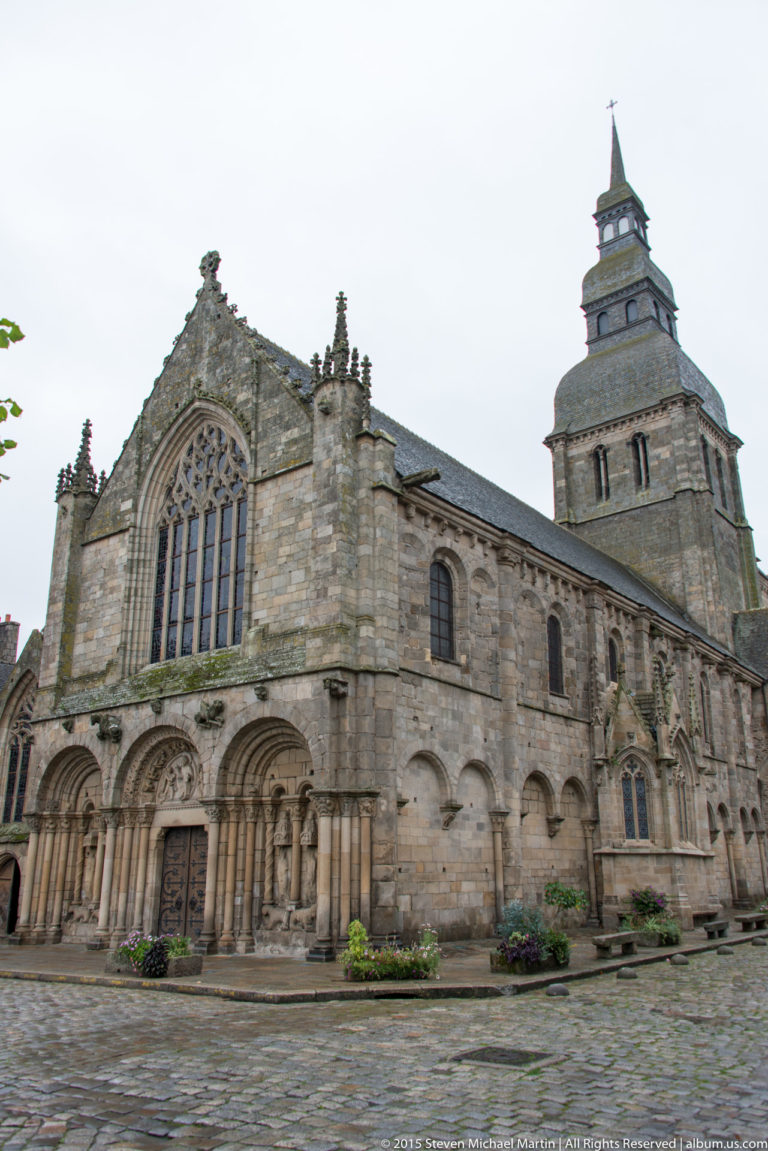 Wide exterior view of the Basilique Saint-Sauveur showing its west façade, long stone side wall, and square bell tower.