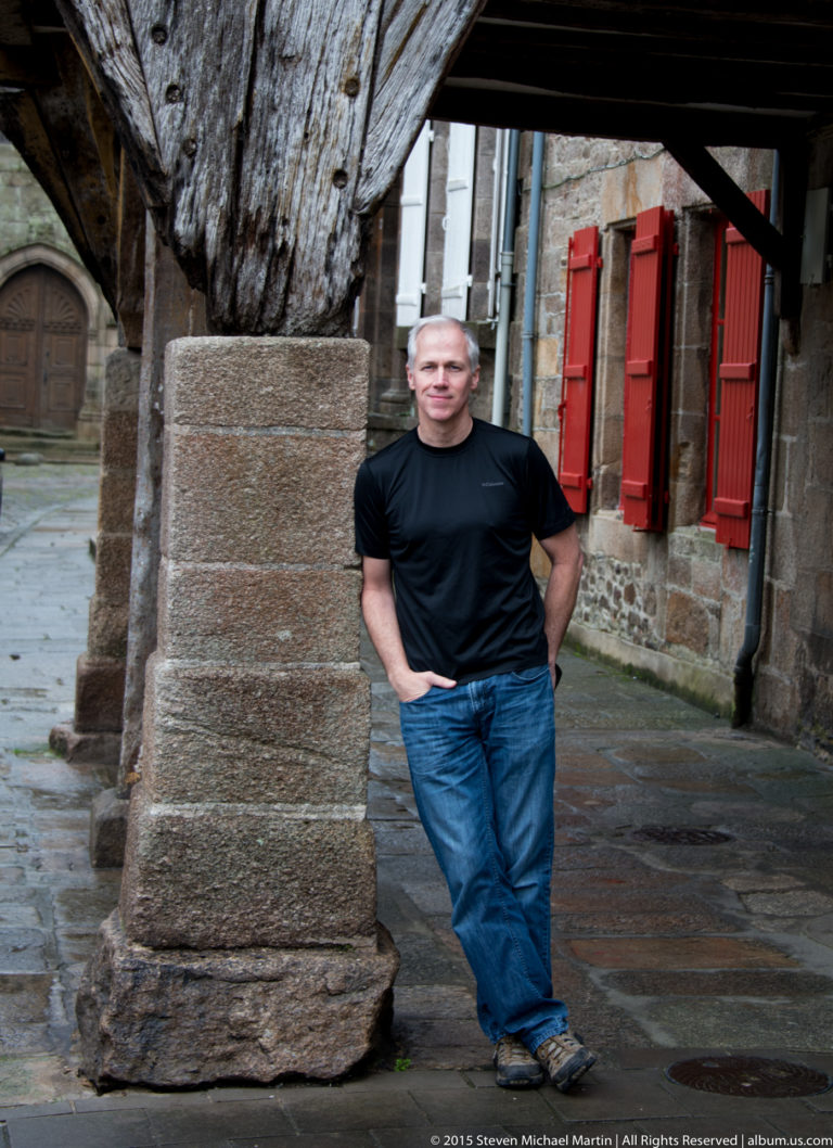 Man in black shirt and jeans leaning beside a massive timber post, with stone walls and red shutters behind him.