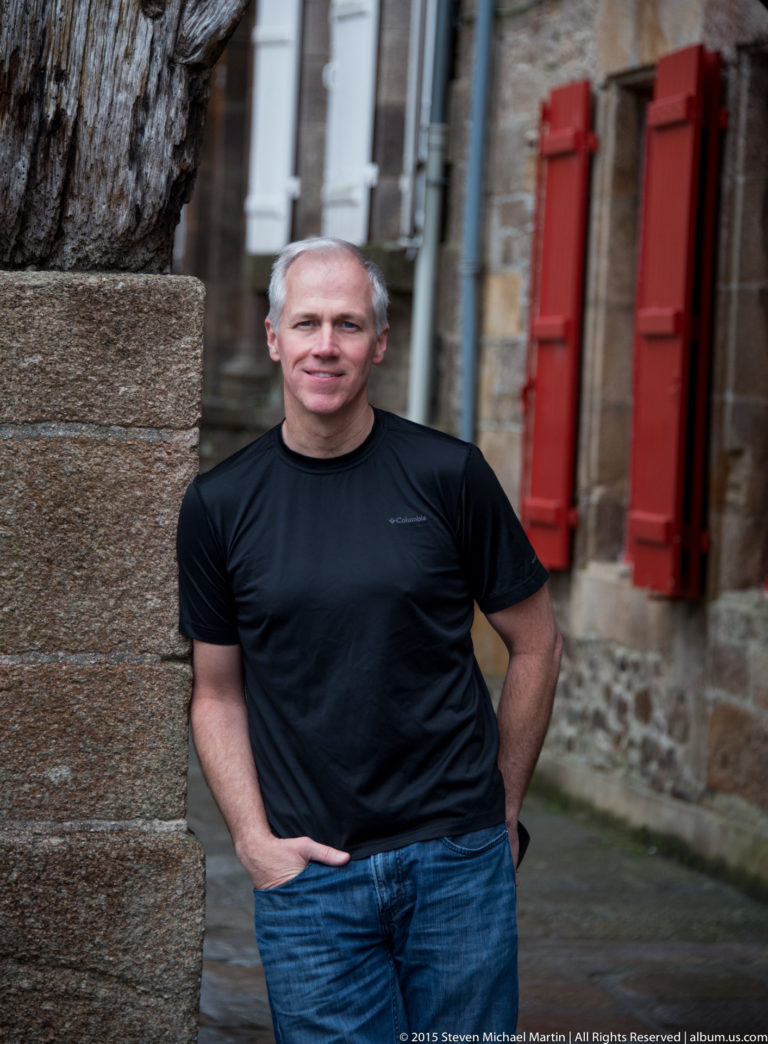 Close portrait of a man in a black shirt standing beside a heavy timber post with red shutters and stone walls behind.