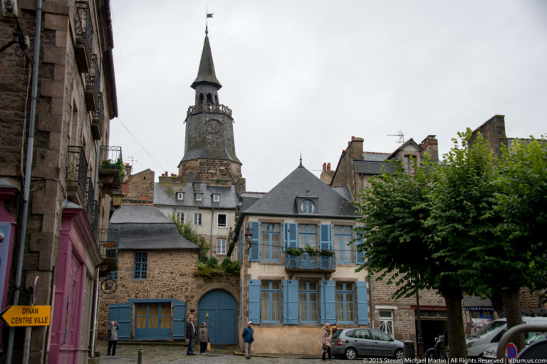Dinan square with historic buildings and blue shutters, pedestrians, trees, and the Tour de l’Horloge rising behind.